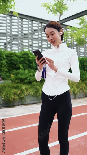Portrait of a smiling Asian sports woman using smartphone with earphones and holding water bottle outdoors. Active healthy lifestyle, music streaming, and wellness concept. Vertical Shot
