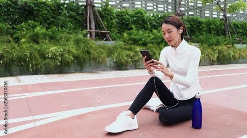 Young Asian woman resting on sports running track with water bottle, using mobile phone with earphones. Active healthy lifestyle, fitness app, hydration, and wellness concept.