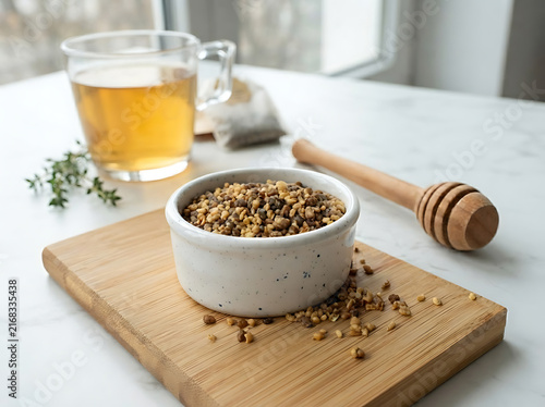 Bowl of raw bee pollen granules with herbal tea and honey dipper on a wooden board.