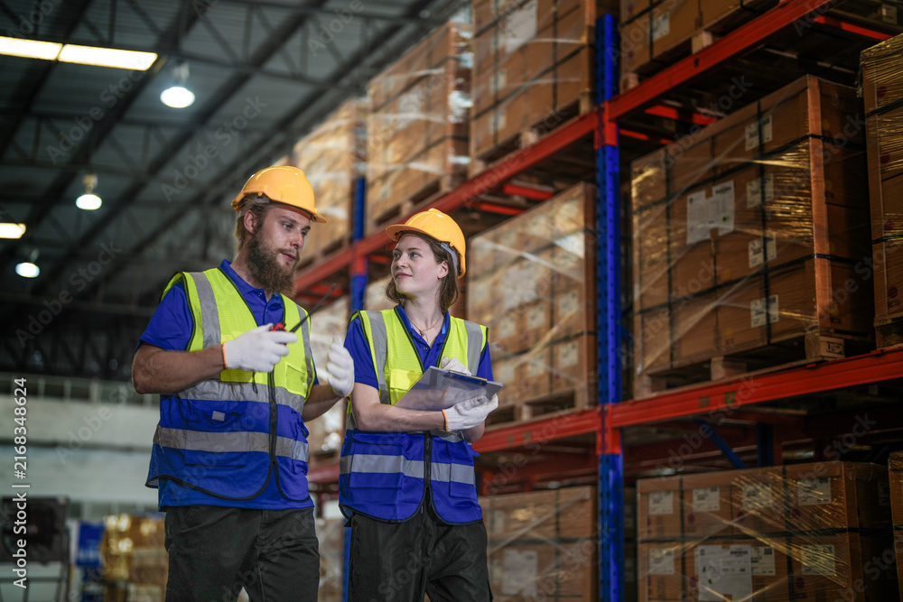 Obraz premium Male warehouse supervisor with a beard and his female coworker in protective workwear hold a two way radio while checking the racks in a large logistics distribution centre with tall.