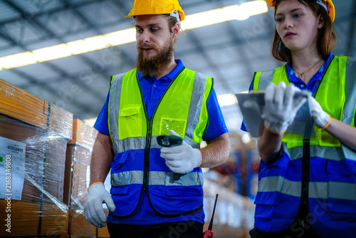 Foto Bearded male warehouse worker and young female colleague in hard hats and safety vests double check package labels against the manifest in a large fulfilment centre with rows of stacked