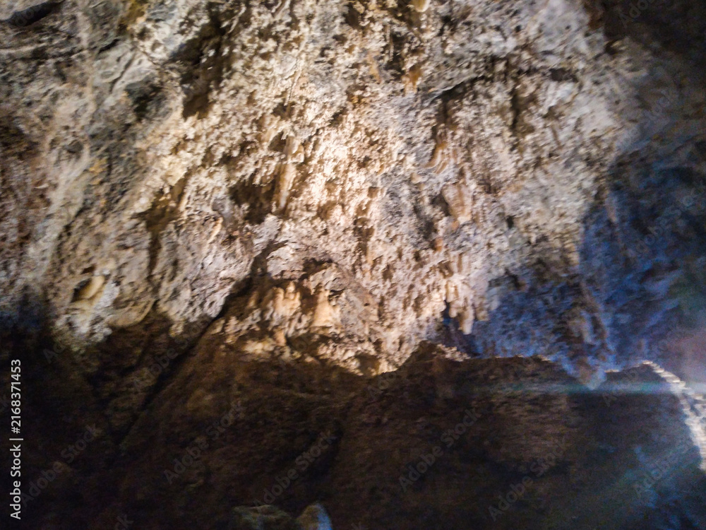 Fototapeta premium Massive limestone cave interior showcasing intricate textured rock formations with natural stalactites hanging from a dark rugged ceiling illuminated by mysterious soft light in underground cavern