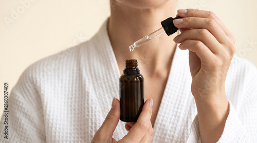 Person in white bathrobe holding amber oil dropper bottle near neck and chest, close up shot on warm background. Dermatological care, body oil packaging and skin hydration concept.