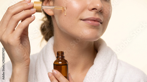 Beautiful woman in plush bathrobe dispensing facial serum dropper onto cheek, close up portrait on beige background. Self-care, moisturizing skincare and facial wellness concept.