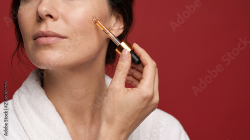 Young woman in white spa robe applying facial serum with dropper on cheek, close up portrait on red background. Facial skincare, anti-aging treatment and beauty cosmetics concept.