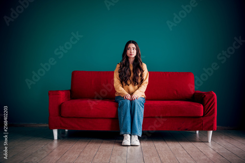 Foto Young woman sitting anxiously on a red sofa against a teal wall, looking worried