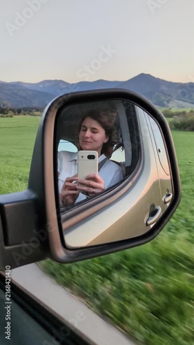 Young woman filming selfie video in car side mirror driving along scenic green meadow and mountains at sunset road trip, joyful female traveler recording vlog memory in moving camper vehicle