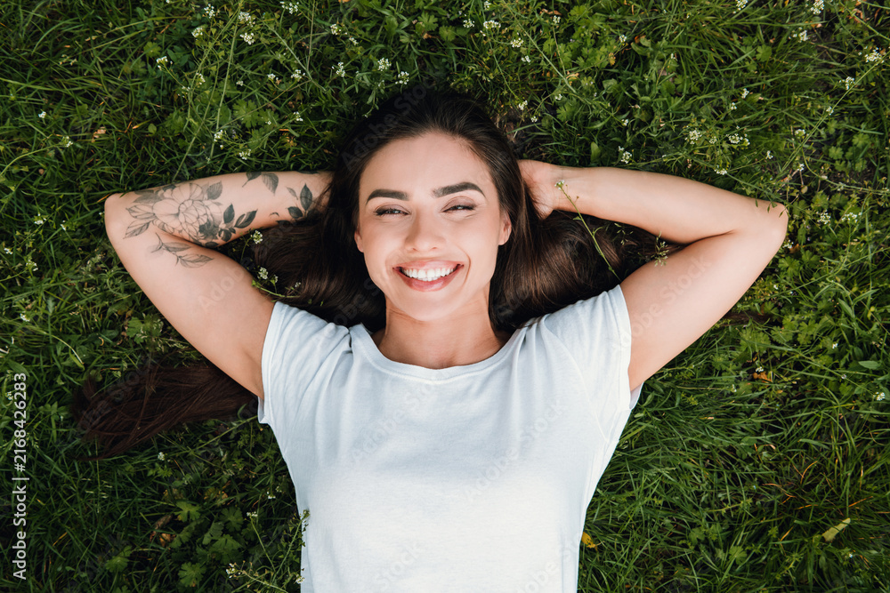 Fototapeta premium Smiling young woman with floral tattoo lying relaxed on green grass outdoors enjoying sunshine and carefree summer relaxation