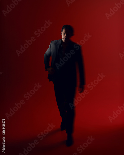 Full length front image of a muscle afro american young man in black suit, keeps his hands in his pockets, looking at the side, isolated on dark red background.