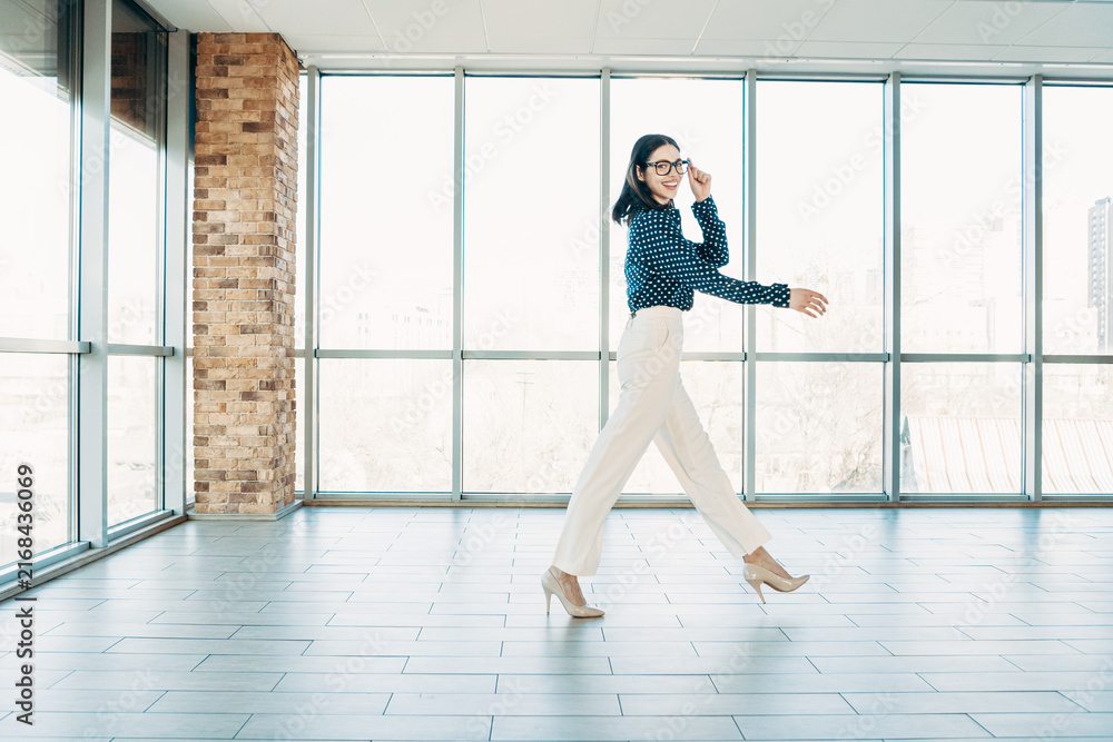 Fototapeta premium Confident businesswoman walking across a bright modern office space in heels and glasses, smiling as she strides with purpose toward a meeting or workspace