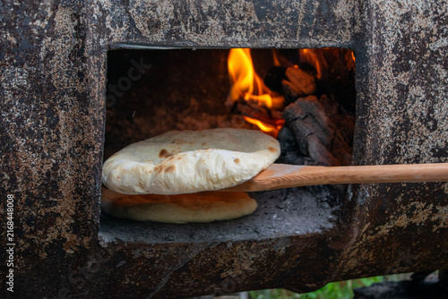 Traditional bread baking inside a rustic wood-fired barrel oven next to burning logs and hot embers. Authentic outdoor baking process in a rural village setting.