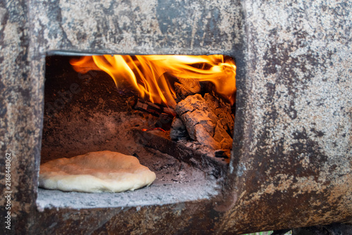 Traditional bread baking inside a rustic wood-fired barrel oven next to burning logs and hot embers. Authentic outdoor baking process in a rural village setting.