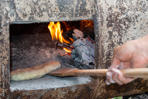 Traditional bread baking inside a rustic wood-fired barrel oven next to burning logs and hot embers. Authentic outdoor baking process in a rural village setting.