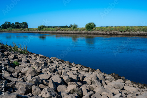 A river under a blue sky with stony banks in a natural environment, dike landscape on the Hunte in summer in 27798 Hude, Germany