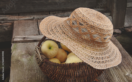 Apples in a basket and a hat on the top. Garden setting. Blurred view on the background. Old wooden floor  background. Home garden fruits, autumnal harvesting, realistic vintage countryside scene