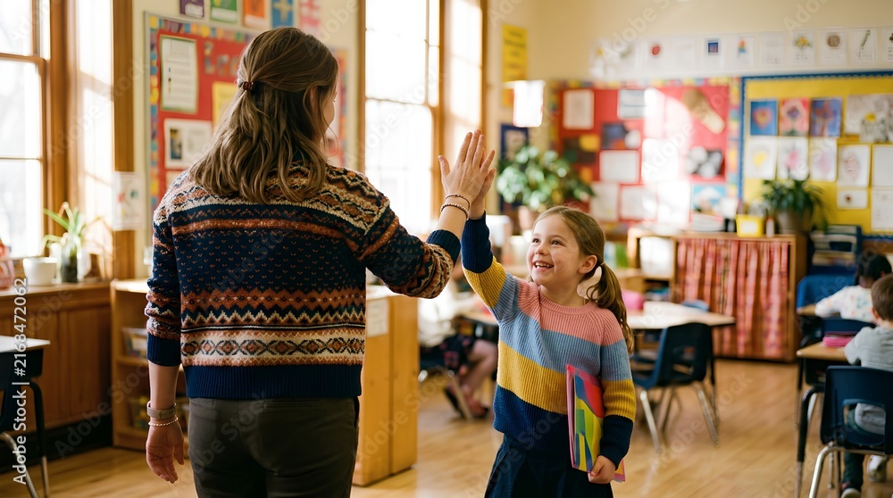 Fototapeta premium Faceless teacher seen from behind giving high-five to smiling young girl in colorful sweater holding rainbow folder in bright, cheerful elementary classroom with artwork and desks
