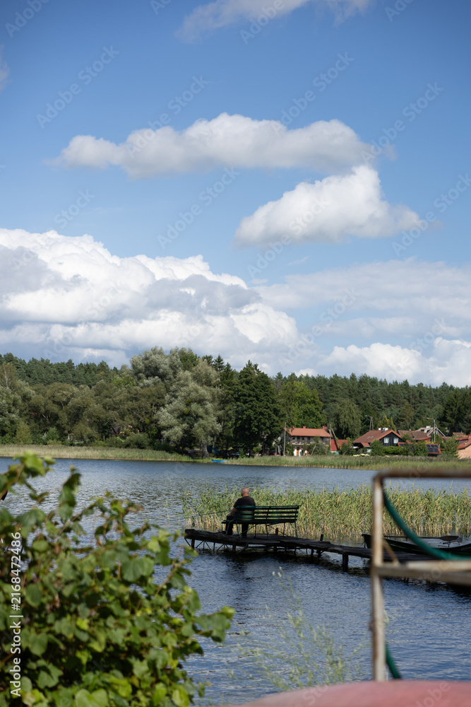 Fototapeta premium Solitary person sitting on a bench at the end of a narrow wooden pier, looking across a peaceful lake toward a rural village under white summer clouds.