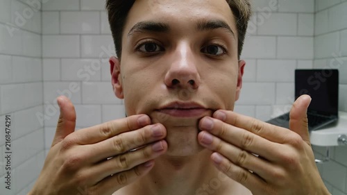 Starting shirtless man massaging face for skincare in tiled bathroom, with laptop on right shelf