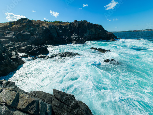 Konstfotografi Waves crash against rocks under a clear blue sky near the coastline during the d