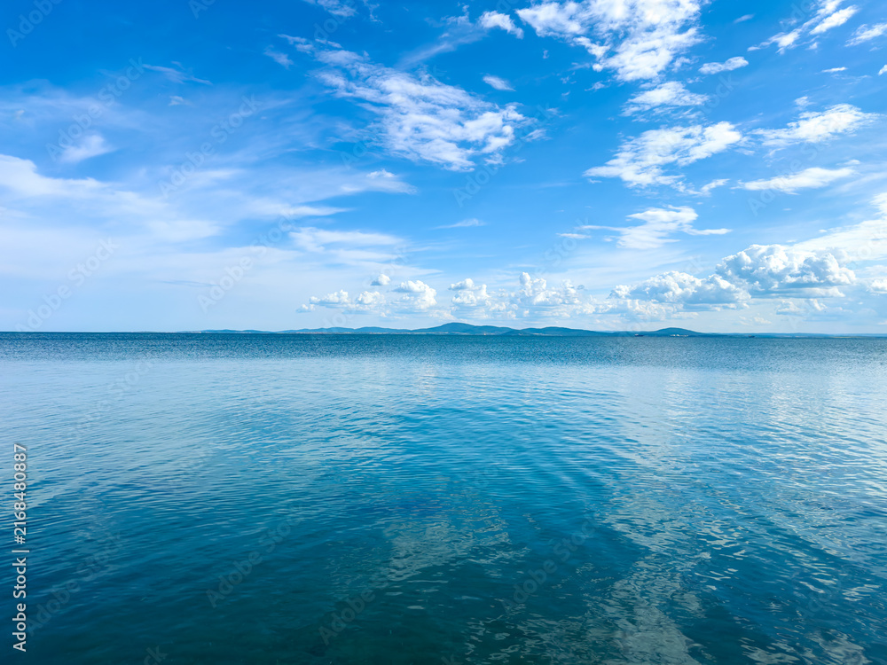 Fototapeta premium Calm water reflects blue sky with clouds over a distant landscape in the early afternoon