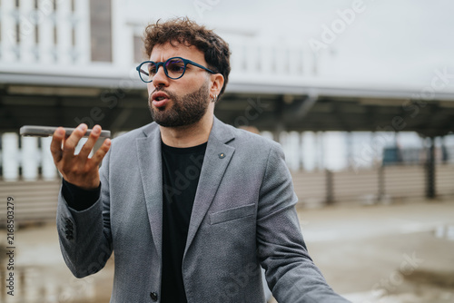 A young businessman speaks into his smartphone holding it at arm’s length. He uses voice messaging or makes a call while standing outdoors in a city environment.