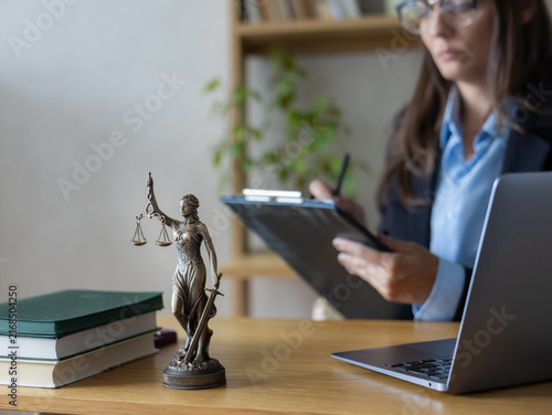 Woman lawyer in glasses using blue smartphone at desk. Bronze Justice statue, green books near open laptop. Focused legal professional handling online client consultations using modern digital tools.