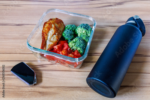 Glass container with roasted chicken breast, broccoli and tomatoes near black water bottle and car key on wooden table, Healthy lunch prep and daily commuting concept