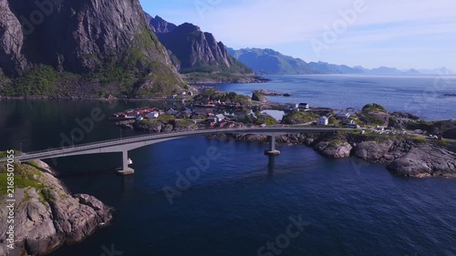 Aerial drone view across the concrete bridge connecting rocky islands and traditional village houses in Lofoten, Norway on a clear sunny day.
