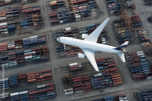 A white airplane is flying over a large, dense container yard in an industrial setting.