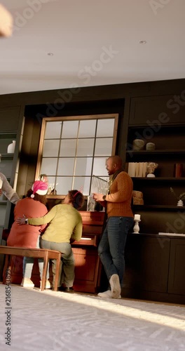 Vertical video: Diverse family gathering by piano at home, person in green reaching keys, Santa hat