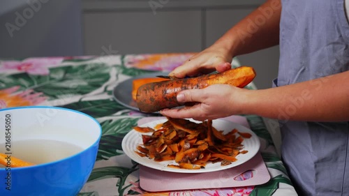 A person is peeling a carrot on a kitchen table with a patterned tablecloth, viewed from a top-down perspective. The scene is set in a well-lit, clean kitchen with a neutral mood, ideal.