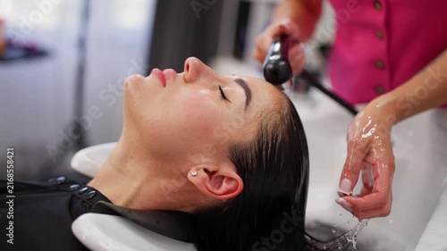 A woman is receiving a hair washing treatment in a relaxed salon setting, with pink and white tones creating a serene atmosphere. The side view captures the sink and the hairdresser at work.