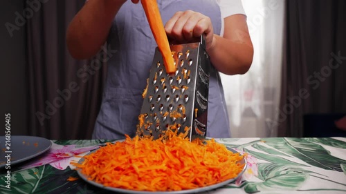 A person is grating carrots using a grater in an indoor setting with curtains and a tablecloth. The scene is captured from a close-up, slightly elevated angle, with a neutral mood.