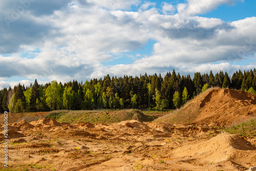 Expansive view of a textured sand quarry featuring piles of loose soil under a dramatic blue sky with fluffy white clouds, bordering a dense coniferous and deciduous forest backdrop