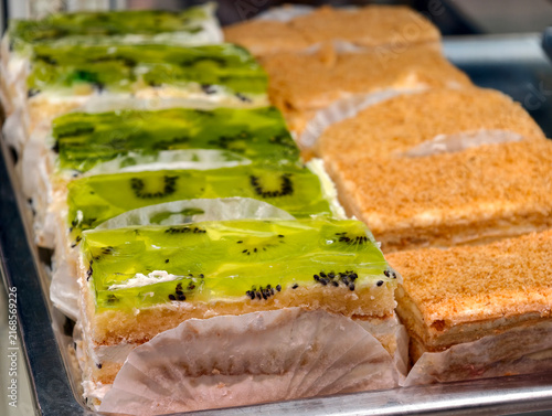 Pastries with green kiwi jelly and pastries decorated with yellow crumbs stand in a confectionery display case, offering a colorful and appealing dessert selection
