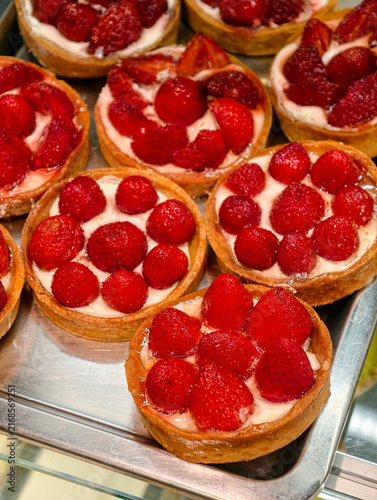 Round tartlets decorated with fresh red strawberries stand on a confectionery display case, creating an elegant presentation of sweet pastries with natural fruit