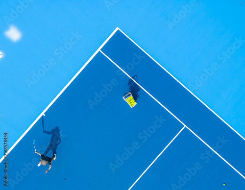 aerial view of a tennis player preparing to serve on a bright blue hard court. Long dramatic shadows, crisp white lines, and a basket of yellow balls create a striking sports composition.