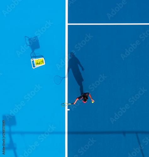 aerial view of a tennis player preparing to serve on a bright blue hard court. Long dramatic shadows, crisp white lines, and a basket of yellow balls create a striking sports composition.