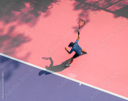 aerial view of a young tennis player in action on a vibrant pink and purple hard court. Dappled tree shadows and strong sunlight create a dynamic and aesthetic summer sports composition.