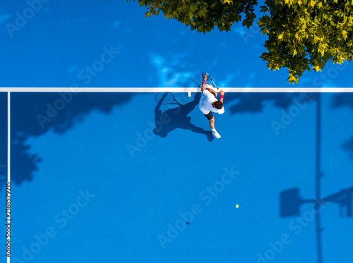 aerial view of a young male tennis player in action on a vibrant blue hard court. Beautiful tree shadows and a sharp silhouette enhance this dynamic outdoor sports composition.