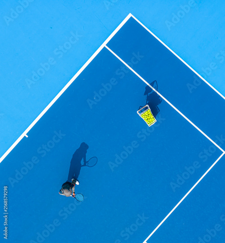 aerial view of a tennis player preparing to serve on a bright blue hard court. Long dramatic shadows, crisp white lines, and a basket of yellow balls create a striking sports composition.