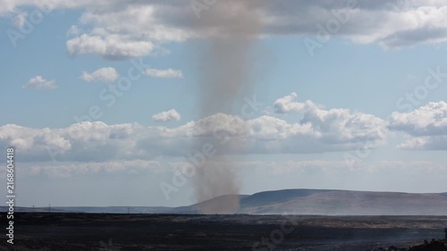 Dust devil formed by intense ground heat spinning over smouldering wildfire aftermath, dark scorched earth stretching toward distant Welsh hills under summer clouds