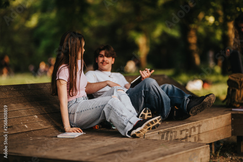 Two students sit and talk on a wooden park bench, wearing casual jeans and t-shirts. They relax outdoors with a notebook nearby, smiling and enjoying a warm afternoon.