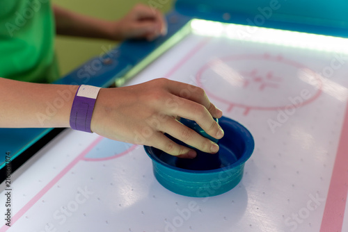 Fotografía Close up of child hand holding blue striker mallet on illuminated air hockey table surface in indoor amusement arcade game room