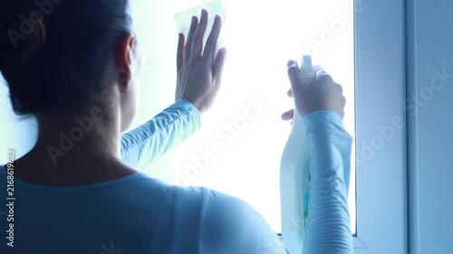A person is cleaning a window in a room using a spray bottle. The focus is on the hand holding the bottle as sunlight comes through the glass.