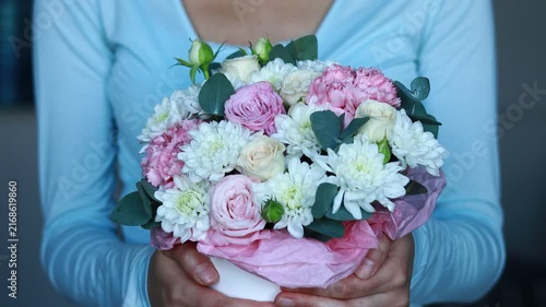 A person stands indoors holding a bouquet of flowers. The bouquet contains pink and white roses, chrysanthemums, and green leaves. The background is blurred.