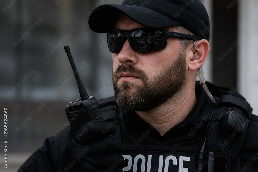 Fototapeta premium A man in a police uniform and cap is pictured outdoors, looking serious while holding a radio.