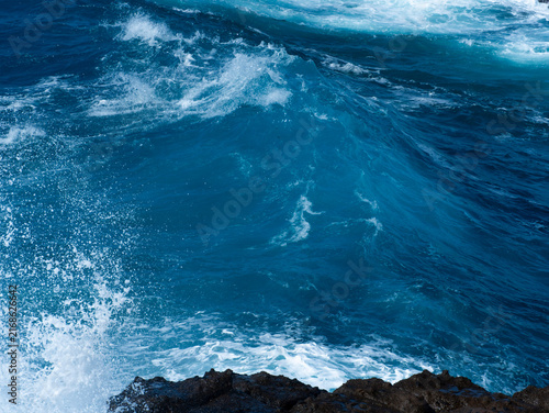 Fotografie Blue ocean waves breaking over dark coastal rocks