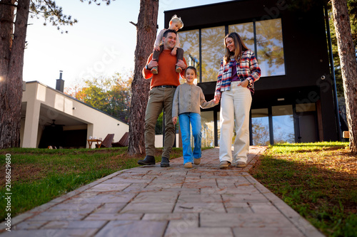 Happy family of four, parents and children, walking down a paved path in front of their contemporary house surrounded by trees, enjoying togetherness and a shared lifestyle