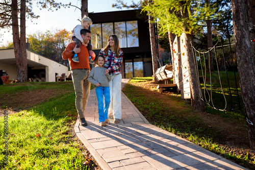 Family with two children enjoying a walk on a sunny day in front of their modern house, with the father carrying his son on his shoulders, symbolizing happiness, childhood, and togetherness